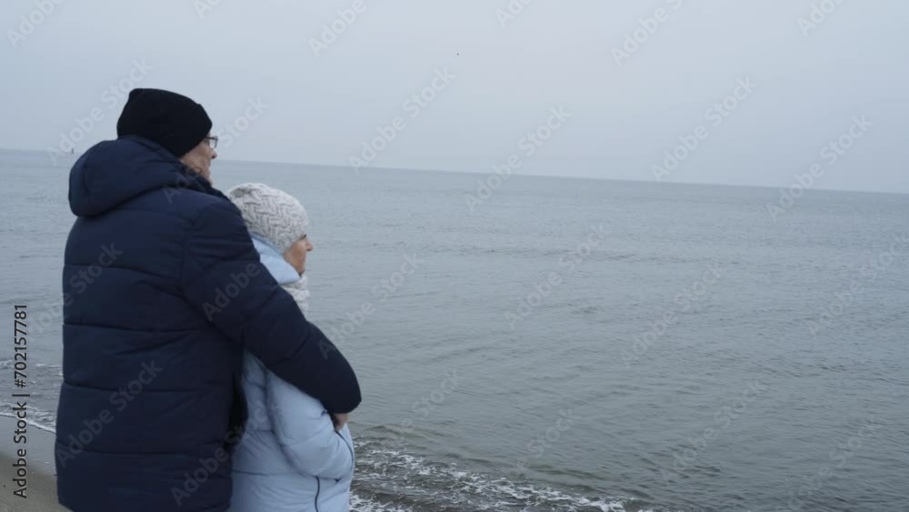 An elderly couple stands hugging on the beach by the sea in winter, looking into the distance. Holidays at sea. An elderly couple stands hugging on the beach by the sea in winter, looking into the distance. Holidays at sea.