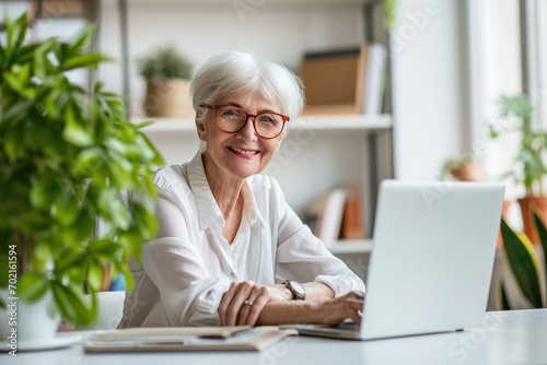 Elderly, Retired woman sitting in white office with laptop, smile, look at camera she is a student studying online with laptop at home, university student studying online