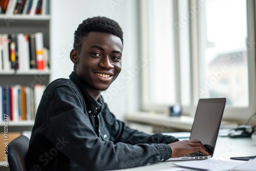 Teenage african man sitting in white office with laptop, smile, look at camera he is a student studying online with laptop at home, university student studying online, online web education concept photo
