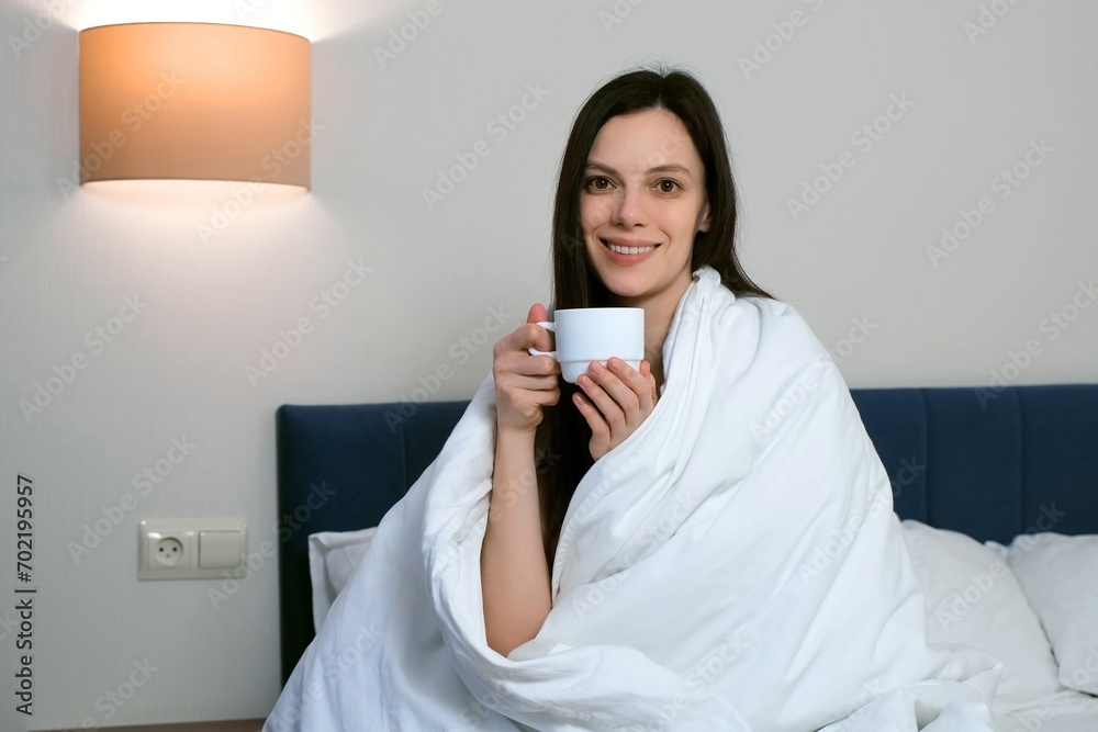 Attractive young brunette woman wrapped in a white blanket smiling with cup of tea or coffee in the morning. She is sitting on the bed and looking at camera. Lazy morning, good mood concept. Attractive young brunette woman wrapped in a white blanket smiling with cup of tea or coffee in the morning. She is sitting on the bed and looking at camera. Lazy morning, good mood concept.