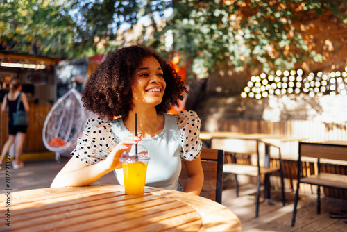 Happy african american teenage girl drinking lemonade or cocktail in an open air cafe. Smiling darkskin female teenager sits at table on the summer terrace. photo