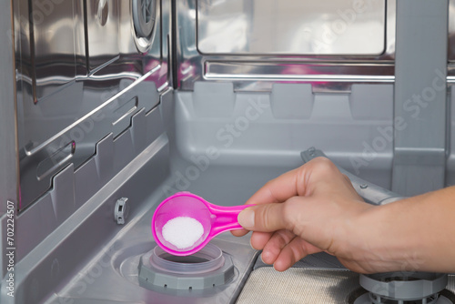 Young adult woman hand holding pink cup or scoop of white salt for dishwasher descaling or water softening. Closeup. Front view.