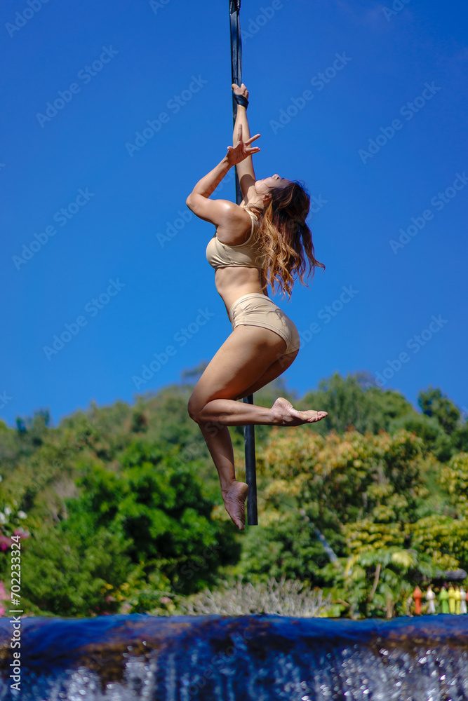 Beautiful Asian girl shows extreme advances by performing pole dance aerial in various positions and spinning stunts on the blue sky around the natural fresh and the waterfall, outdoors.