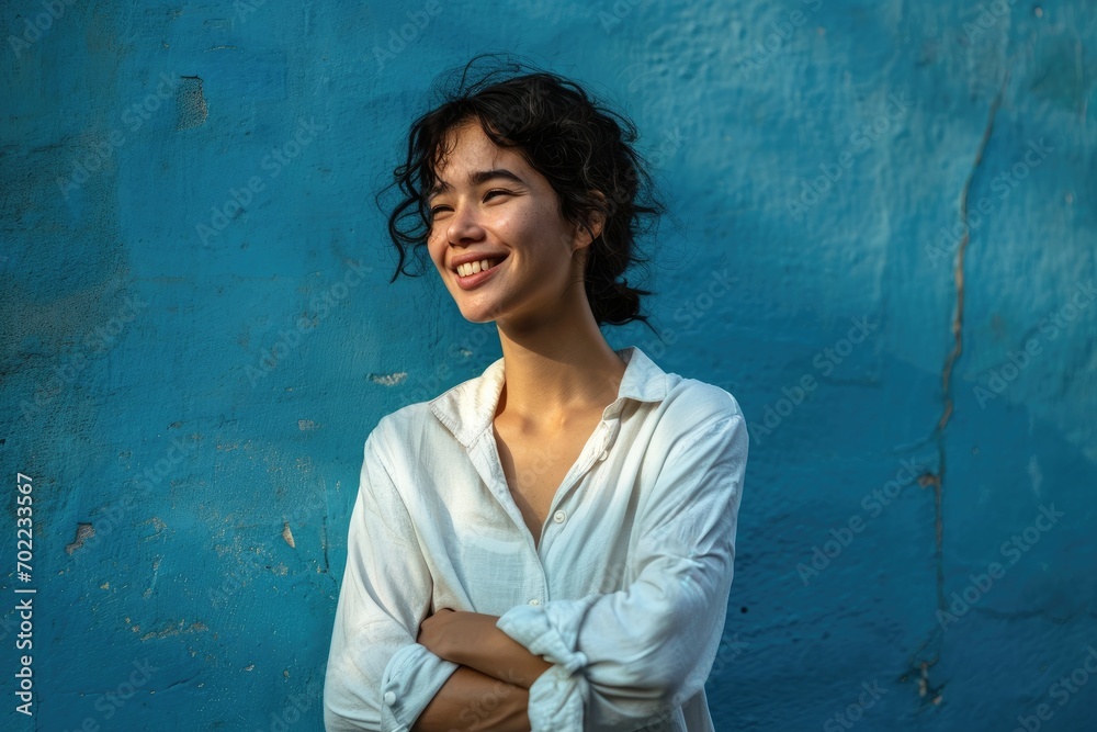 A confident woman stands before a vibrant blue wall, radiating joy as she crosses her arms and flashes a warm smile A confident woman stands before a vibrant blue wall, radiating joy as she crosses her arms and flashes a warm smile