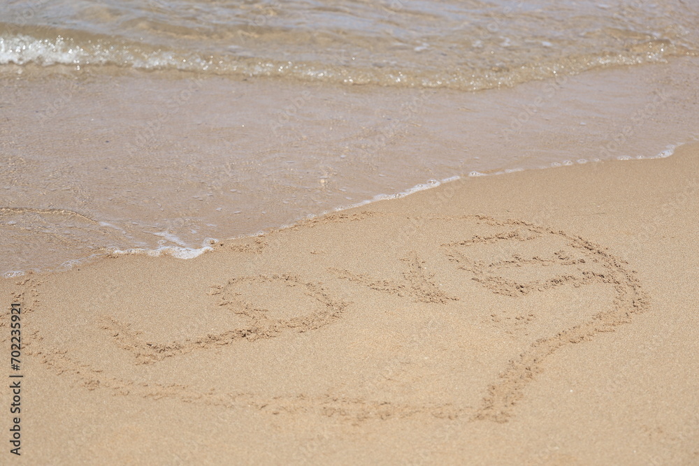 Written LOVE surrounded by a heart shape on a fine sand background. Handwritten on the wet beach sand with a small wave lapping.

