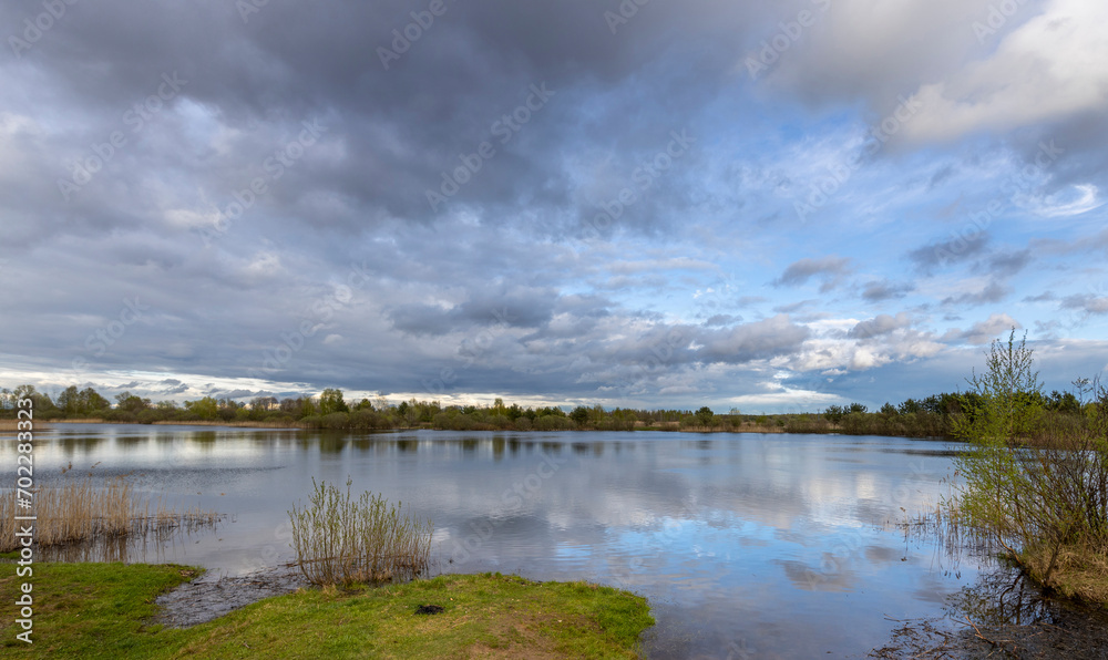 Dramatic spring landscape with clouds reflected in a pond. bank with green grass in the foreground