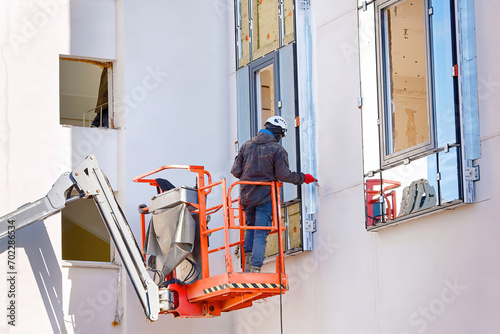 Man in lift bucket caulk, seal exterior window with putty knife, maintain exterior window. Worker in lifting platform at height insulate and seal around building windows. Builder in crane basket photo