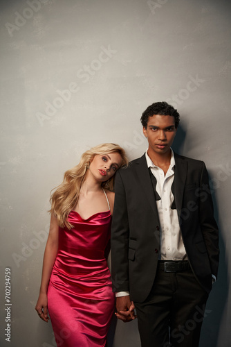 appealing young multicultural couple in elegant evening attires posing together on gray backdrop