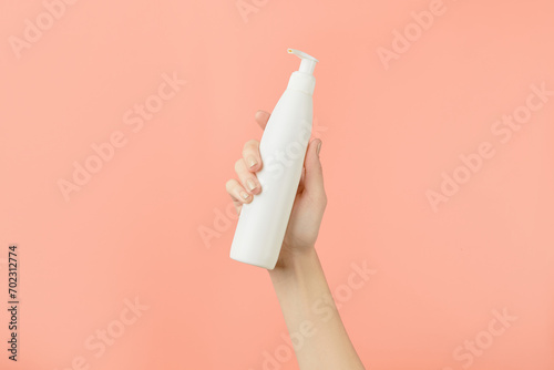 A womans hand holding a white cosmetic bottle on a peach background. Beauty treatment concept photo
