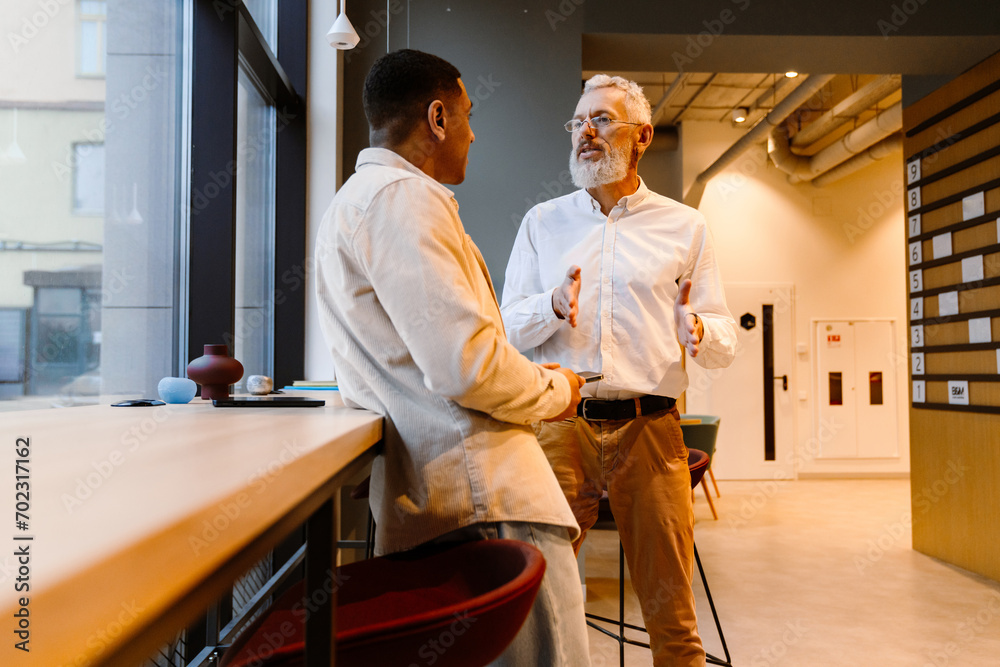 Confident businessman talking to younger male colleague while standing in office space Confident businessman talking to younger male colleague while standing in office space