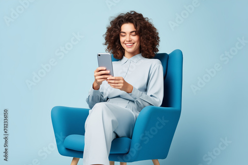 happy girl in sweater pants sitting on blue armchair look at smartphone isolated on pastel color background