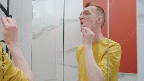Adult man shave off beard on face in bathroom. Shaving stubble with white foam and disposable razor. Skincare routine, careful face washing. Handsome male in casual clothes looking in mirror.
