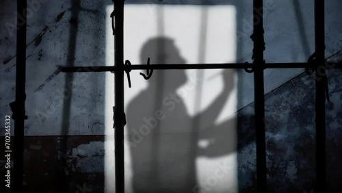 From the cracks on the gate of a prison cell, sunlight projecting a shadow on a worn-out wall: a man painting in the air; symbolic shot, freedom, art, escape.
