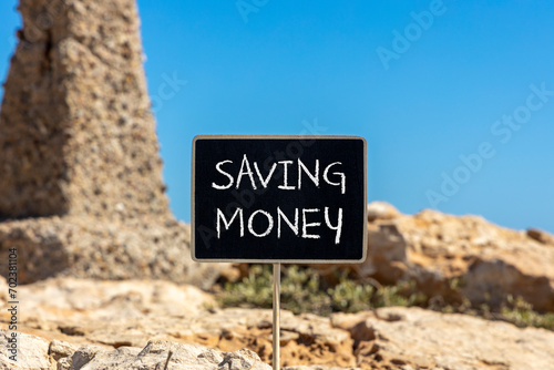 Saving money symbol. Concept words Saving money on beautiful black chalk blackboard on a beautiful beach stone blue sky background. Business, support and saving money concept. Copy space.
