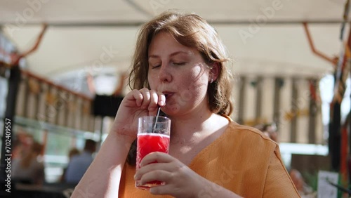 Happy plus size woman stirs berry smoothie and drinks it on sunny terrace in cafe. Portrait of lady drinking soft drink on hot summer day. Charming woman with freckles on plump cheeks. photo