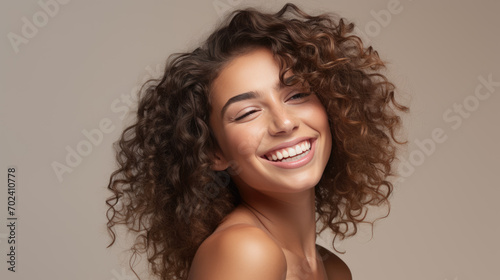 Woman with voluminous curly hair and a beaming smile  wearing a tank top and posing against a warm beige background.