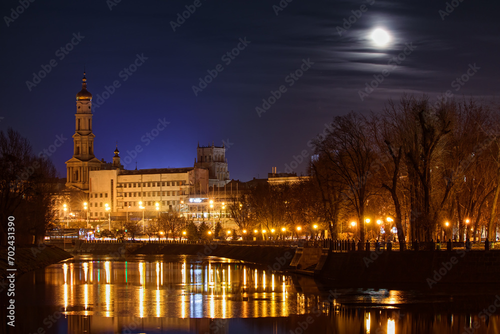 Fototapeta premium The Lopan River with an embankment and a park in Kharkiv city (Ukraine). Evening cityscape of the city center with buildings and a Christian church