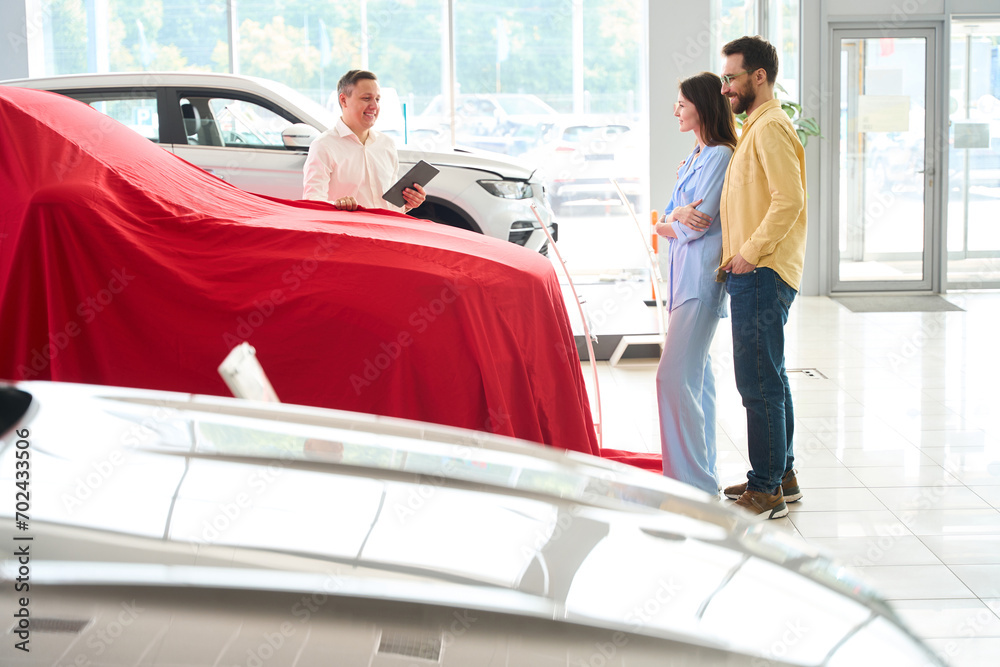 Car dealership consultant presents a car under a red cover Car dealership consultant presents a car under a red cover