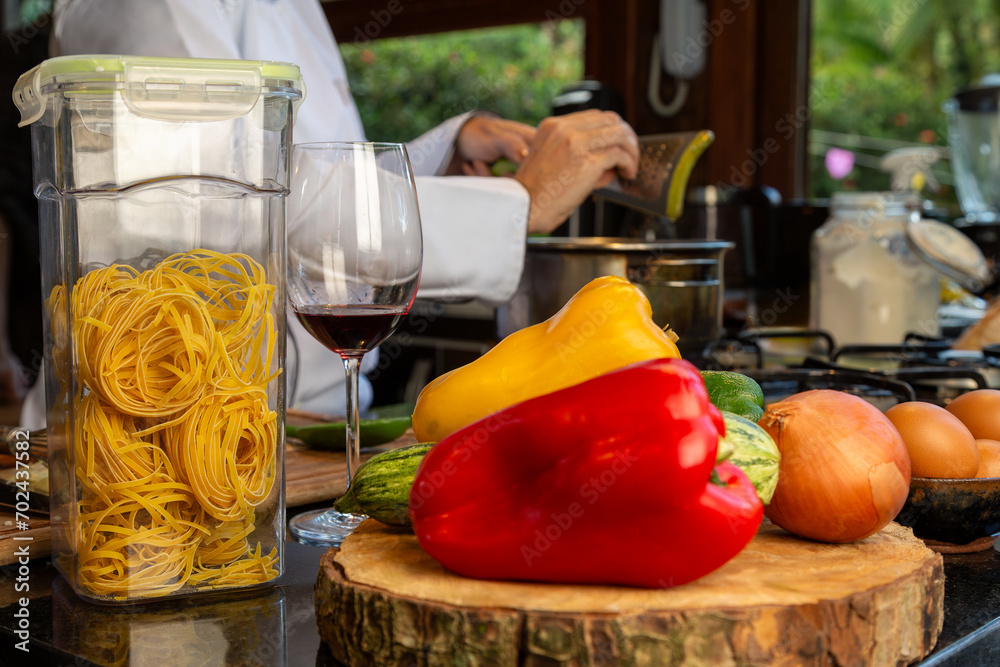 Obraz premium Bancada de cozinha com pimentões amarelo e vermelho, cebola e abobrinha sobre uma tábua de tronco, um pote de massa de talharim crua, uma taça de vinho e chef de cozinhando ao fundo.