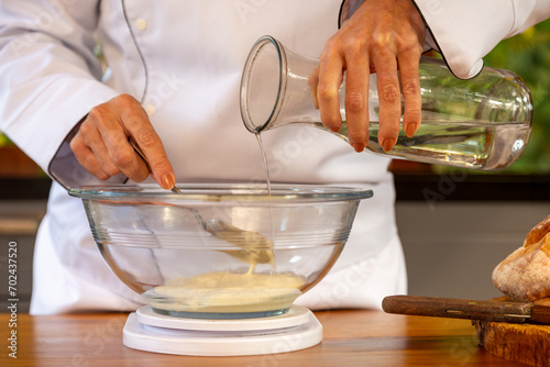 Chef de cozinha mulher em bancada de madeira, segurando uma garrafa de vidro despejando água numa tigela de vidro com farinha em cima de uma balança, no preparo para uma massa de pão. photo