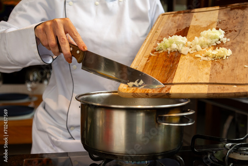 Chef de cozinha com dólmã branco segurando tábua de corte em uma mão e na outra um facão inox, enquanto joga na panela inox os temperos de alho e cebola que estão na tábua.  photo