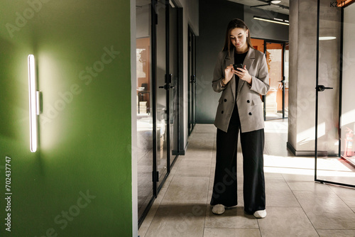 Stylish businesswoman texting on smartphone in a modern office corridor, bathed in natural light.