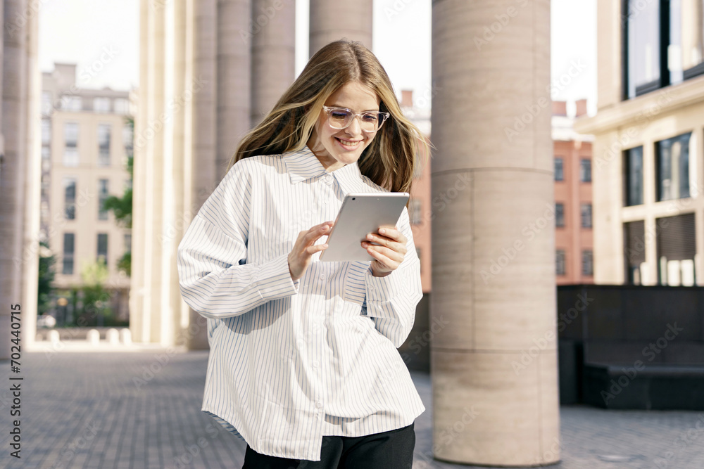 Joyful young woman using a tablet on the go, with stylish eyewear and modern city architecture in the background. Joyful young woman using a tablet on the go, with stylish eyewear and modern city architecture in the background.