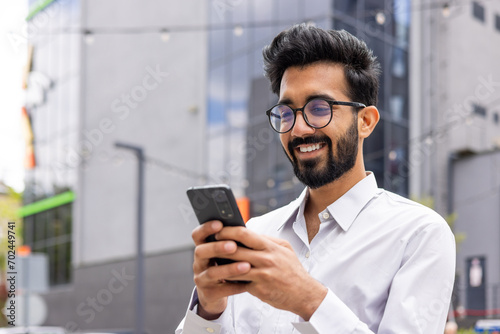 Young Hindus Businessman walks through a city with a phone in his hands, a man in business clothes is satisfied with a smiling reading message and viewing social networks, outside office.