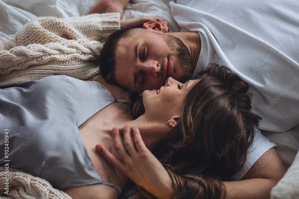 Beautiful happy loving young smiling couple relaxing in bed, looking at each other. Cozy home atmosphere, tenderness, closeness. Embracing, kissing. Lazy weekend, slow living concept Beautiful happy loving young smiling couple relaxing in bed, looking at each other. Cozy home atmosphere, tenderness, closeness. Embracing, kissing. Lazy weekend, slow living concept