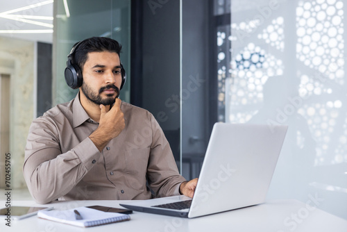 A serious thinking Hindus works inside an office with a laptop, a man in headphones listens to podcasts and audio books, a focused programmer encodes new software. photo