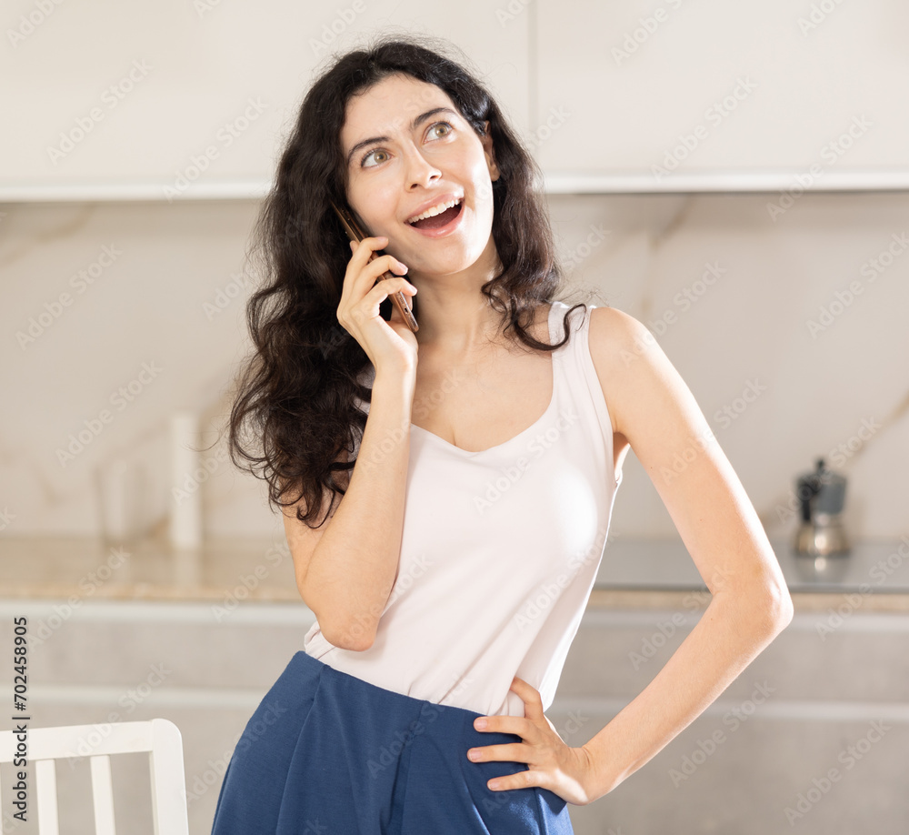 Fototapeta premium Joyful woman talking on smartphone while sitting on kitchen table