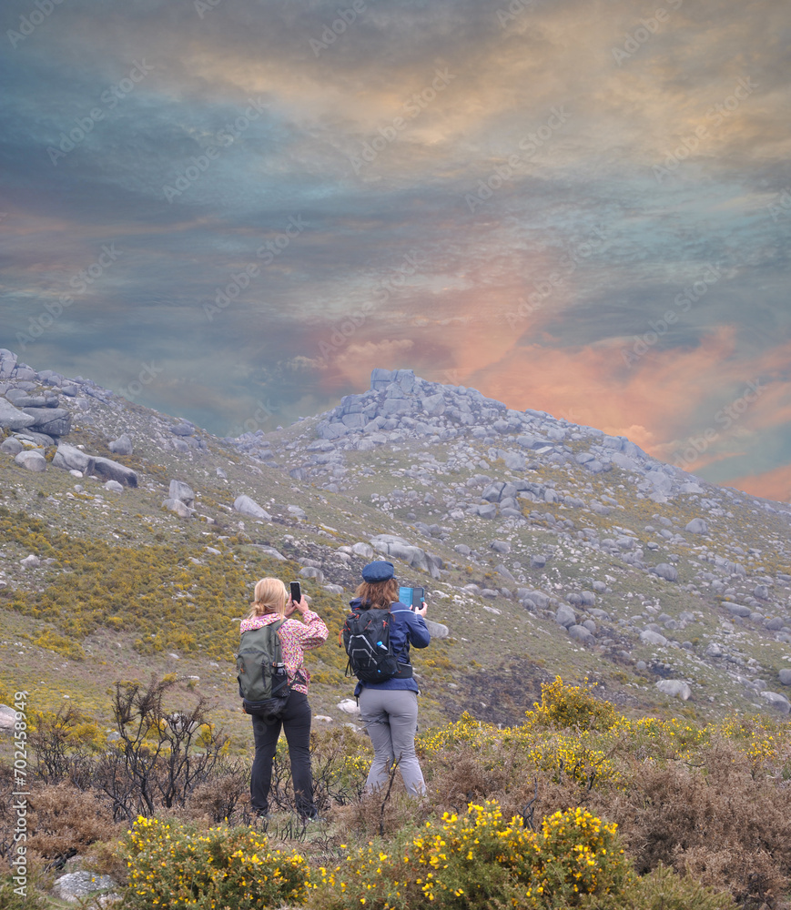 Two women walking on a mountain trail, stopping to observe and photograph the landscape, enjoying nature