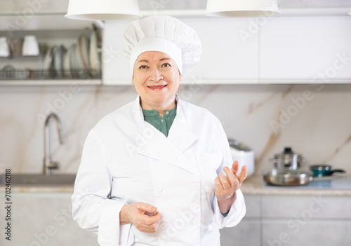 Happy mature female cook in uniform standing and showing her kitchen