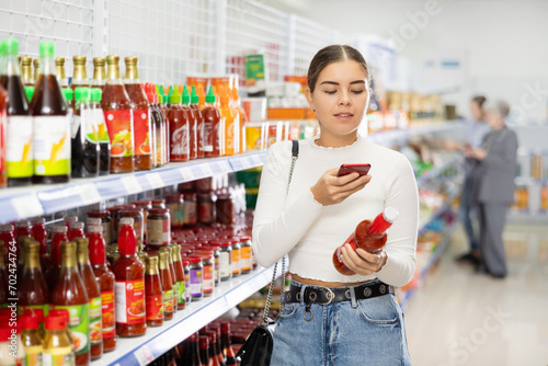 Girl customer in store chooses and take photo of bottle with red sriracha sauce for cooking traditional oriental dishes. Buyers examines package with oriental seasoning