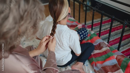 Back view of gray haired young mom braiding her dyed hair daughters pigtail at home in morning