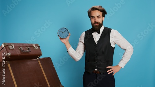Hotel employee checking time on watch, working as bellboy or doorkeeper and holding wall clock to look at hour. Valet in formal clothing posing next to trolley bags, trying to be punctual. photo