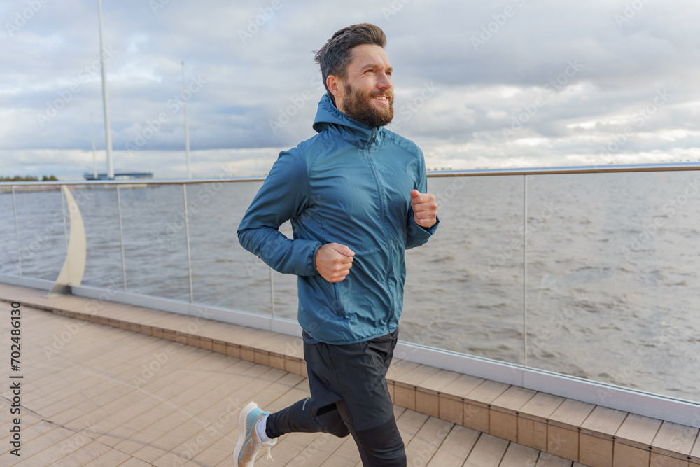 Fototapeta premium Focused man in sportswear running along the waterfront, with urban skyline in the background
