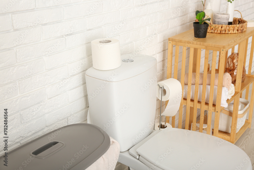 Roll of paper on toilet bowl and rack with bath supplies near light brick wall, closeup Roll of paper on toilet bowl and rack with bath supplies near light brick wall, closeup