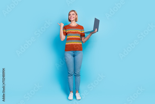 Full length photo of cute girl wear strited t-shirt holding laptop directing look at offer empty space isolated on blue color background photo