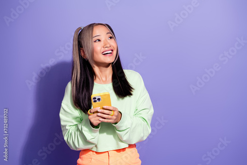 Photo of youth girl with ponytails wear green pullover hold smartphone look right corner empty space isolated on purple color background