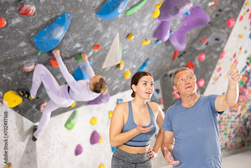 Positive friendly young female coach of climbing gym instructing interested aged male beginner while standing together against backdrop on colored artificial bouldering wall.