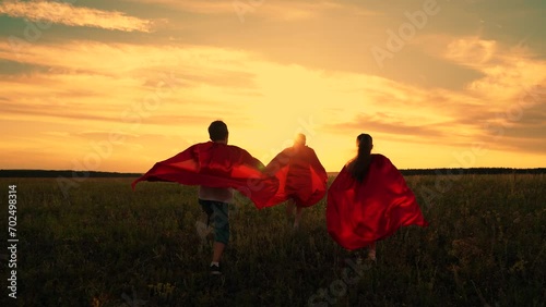 Child imagine being comic book heroes running across meadow in red capes. Children in red capes run across field pretending to be superheroes. Kids actively spend evening dressed as superheroes, dream photo