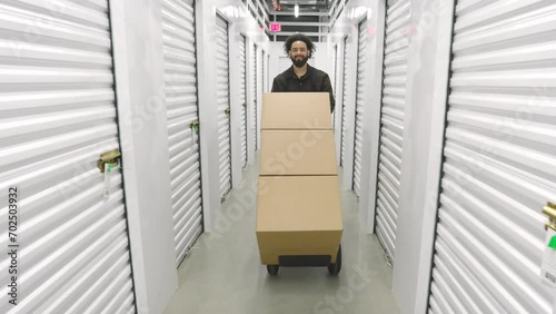 Workday of young male Afro-American in climate-controlled storage, dark-haired man with a trolley cart transporting goods in big boxes, white metal doors. High quality 4k footage