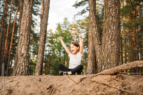 Happy female kid with open mouth sitting on ground in forest with raised arms. Sporty girl enjoying hiking in nature.