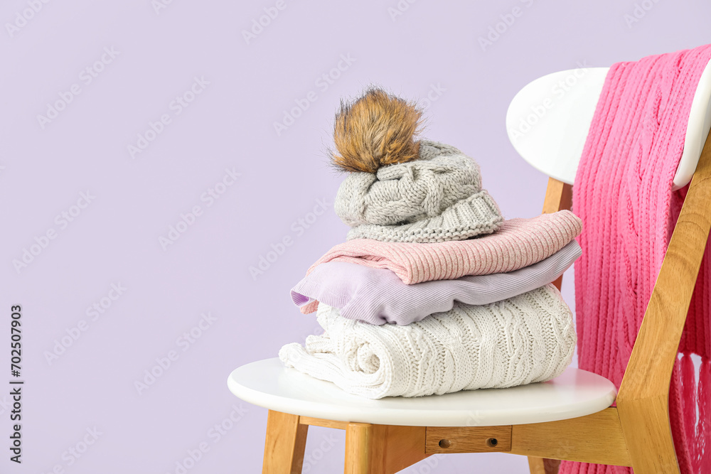 Stacked sweaters with hat and scarf on chair against lilac background Stacked sweaters with hat and scarf on chair against lilac background