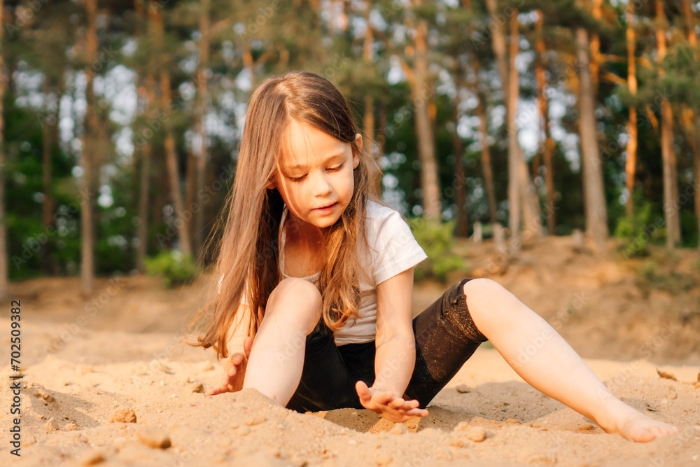 Little girl with dark hair sit on sand near forest. Female child in white t-shirt and black shorts have fun in nature.