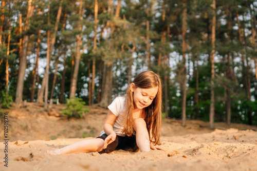 Little girl with dark hair sit on sand near forest. Female child in white t-shirt and black shorts have fun in nature.