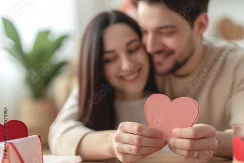 Two young lovers share a special moment holding a pink cardboard heart and roses on Valentine’s Day In an intimate celebration in the living room at home