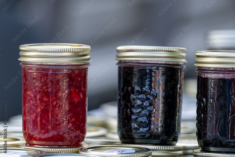 Multiple glass jars of various homemade strawberry, raspberry, and blueberry jam for sale at a market. The crocks have metal lids. The small pureed jelly bottles are made with organic berries. Multiple glass jars of various homemade strawberry, raspberry, and blueberry jam for sale at a market. The crocks have metal lids. The small pureed jelly bottles are made with organic berries.