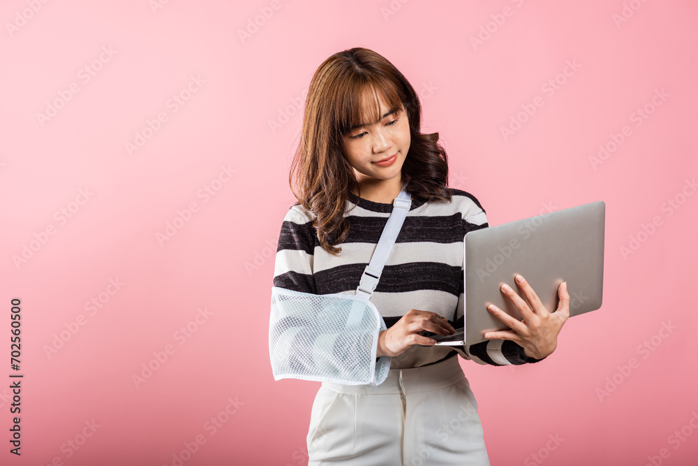 Fototapeta premium An Asian businesswoman smiles confidently with her broken arm in an arm splint. She remains dedicated to work, holding a laptop in a studio shot on a pink background.